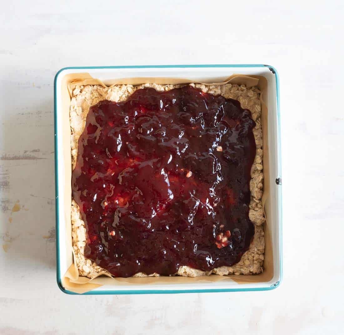 A square baking pan lined with parchment paper contains an oatmeal crust topped with a layer of glossy, red berry jam. The pan is placed on a light-colored surface.