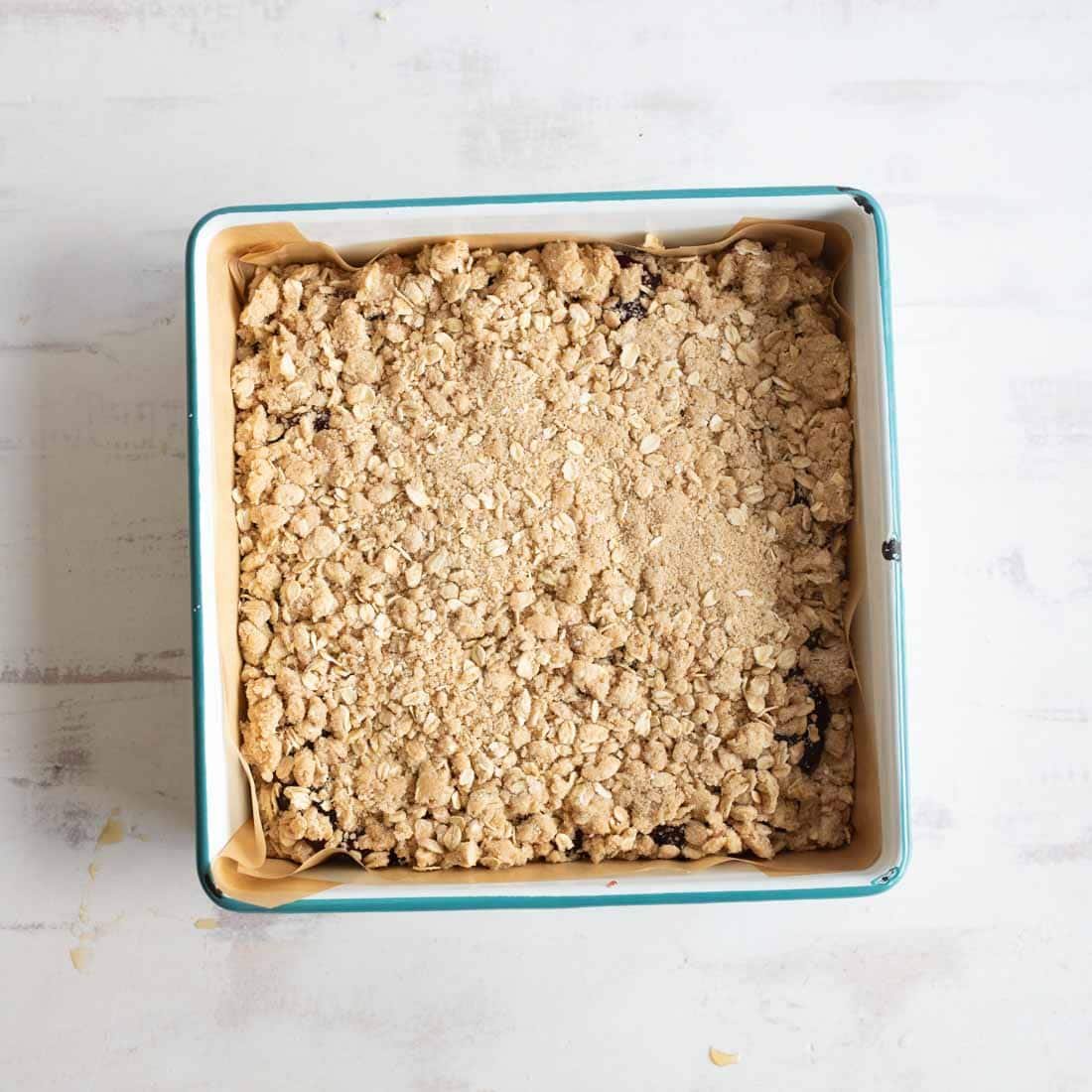 A square baking pan lined with parchment paper containing an unbaked oat crumble topping, evenly spread, ready to be baked.