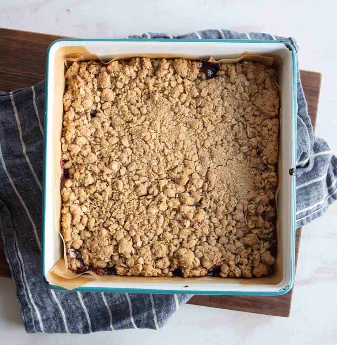A square baking dish filled with golden-brown crumble bars sits lined with parchment paper atop a blue-and-white striped kitchen towel on a wooden board.