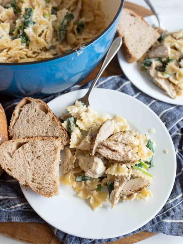 A plate of bowtie pasta in creamy garlic parmesan chicken and pasta sauce with spinach and sliced chicken is served with two slices of bread. In the background, there’s a pot of pasta and another plate with bread.