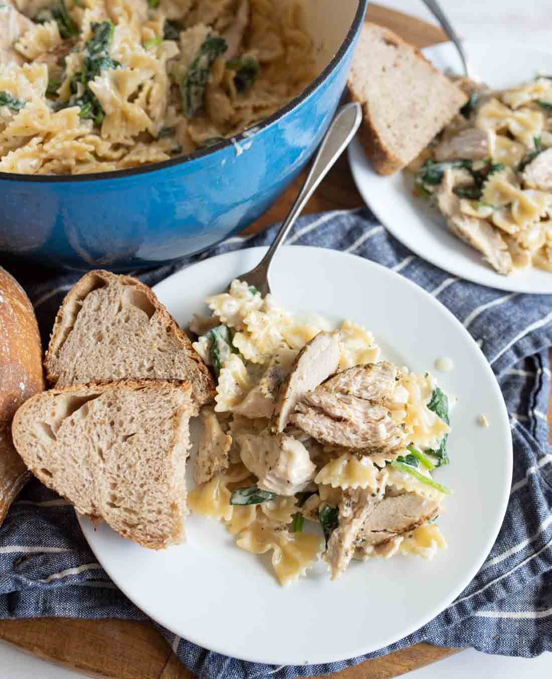 A plate of creamy bow-tie pasta with spinach and grilled chicken slices, served with two slices of rustic bread. In the background, a pot with more pasta and a partial view of another plate are visible.