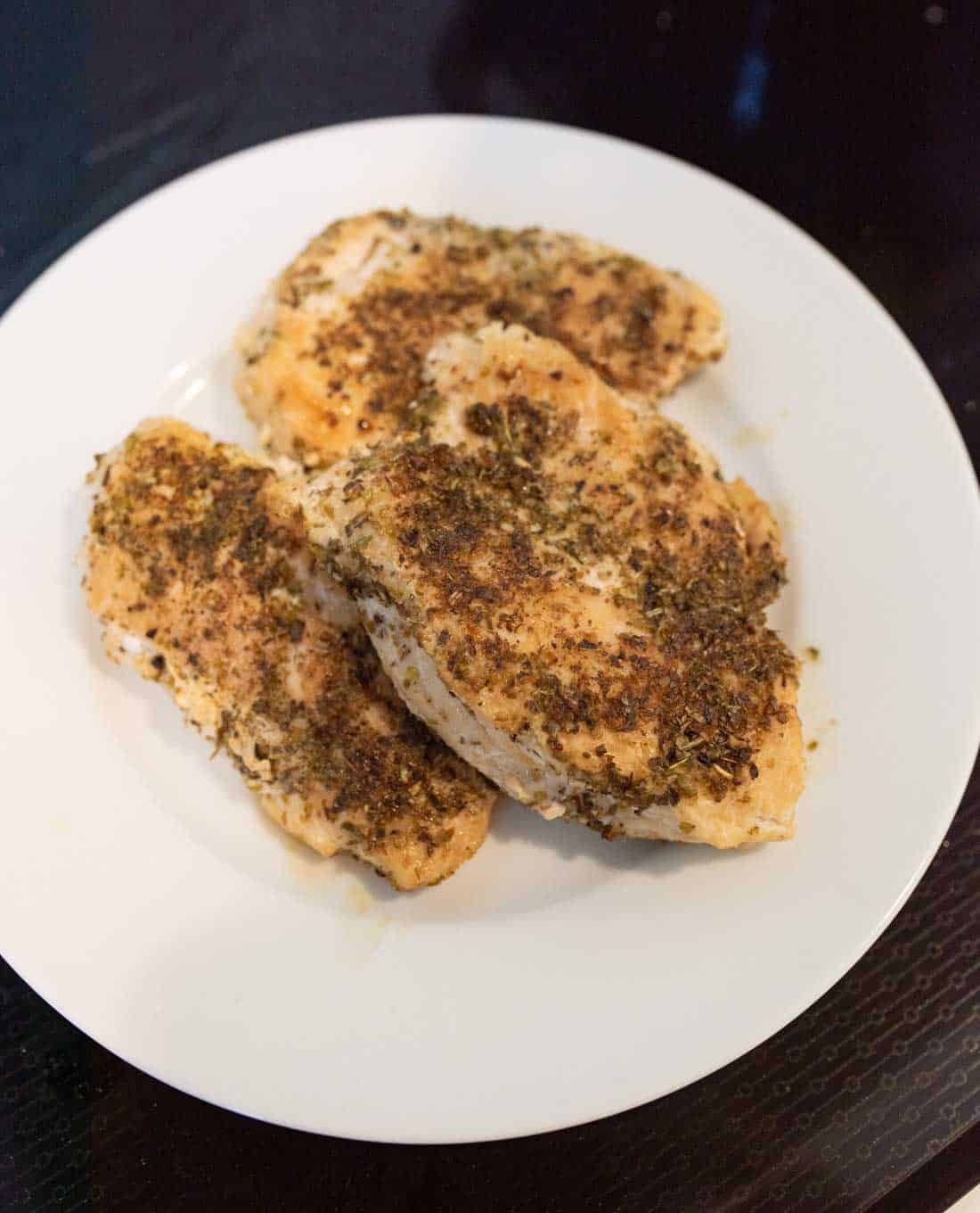 Three pieces of seasoned, cooked chicken breast are arranged on a white plate, displayed on a dark surface. The chicken is coated with a blend of herbs and spices.