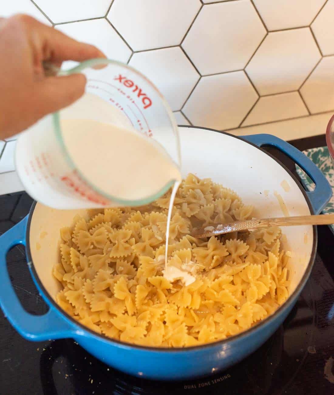 A hand pours milk from a Pyrex measuring cup into a blue pot filled with cooked bowtie pasta on a stovetop, with a fork stirring the pasta. White hexagon tiles are in the background.
