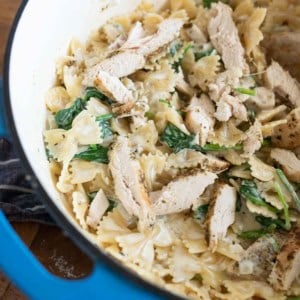 A close-up of creamy bowtie pasta with spinach and sliced grilled chicken in a large blue-edged pot on a wooden surface.