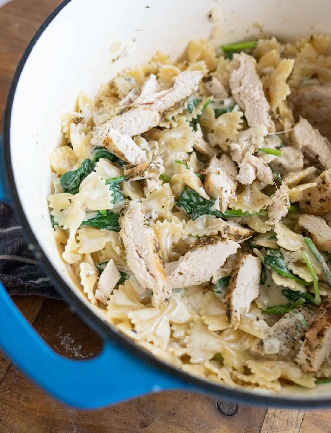 A close-up of creamy bowtie pasta with spinach and sliced grilled chicken in a large blue-edged pot on a wooden surface.