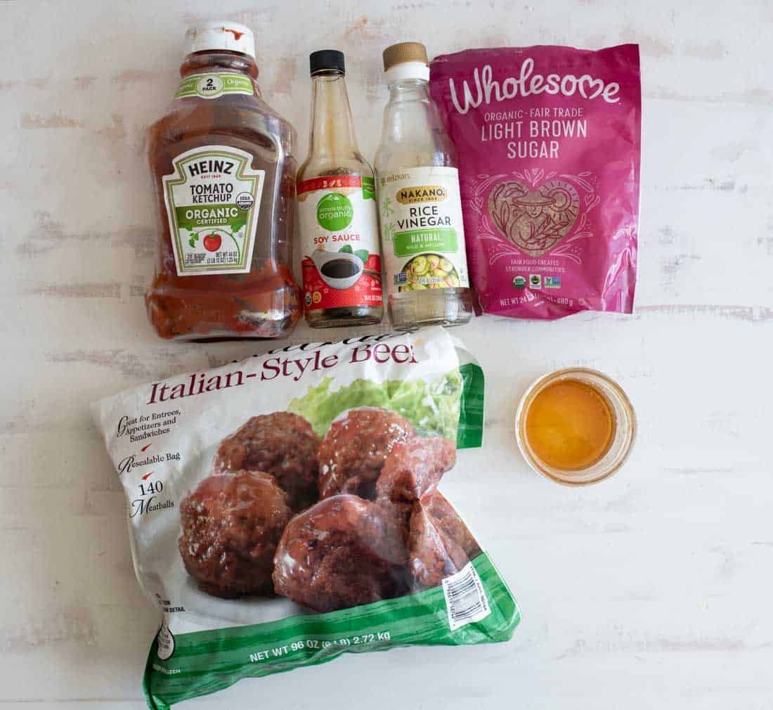 A bottle of ketchup, soy sauce, rice vinegar, a bag of light brown sugar, frozen Italian-style meatballs, and a small bowl of sauce are arranged on a white surface.
