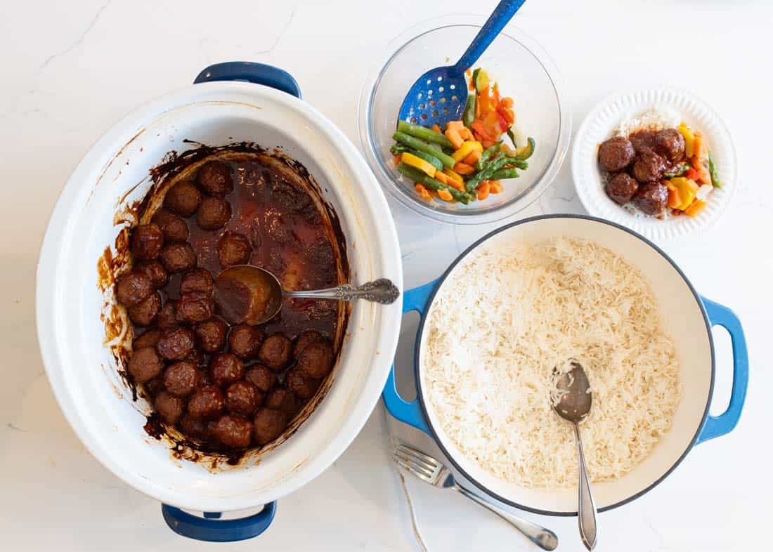 A table with a slow cooker of meatballs in sauce, a pot of cooked white rice, a bowl of mixed vegetables, and a small plate with a serving of rice, meatballs, and vegetables. Serving utensils are nearby.