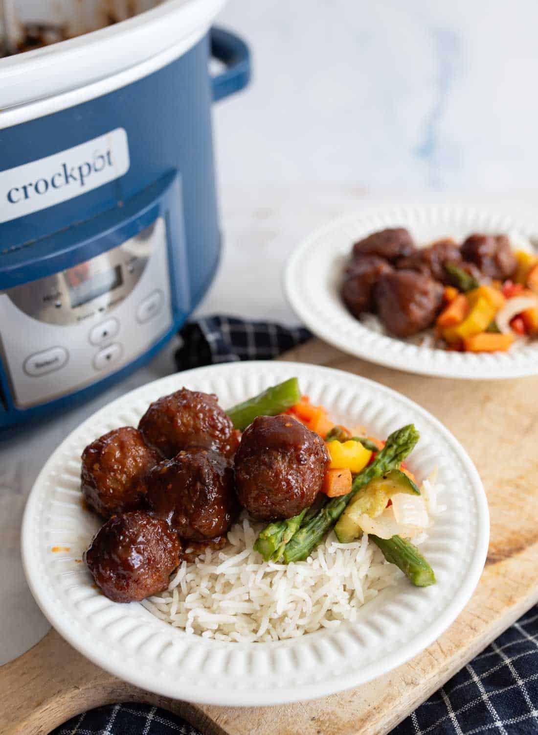A plate with rice, glazed meatballs, and mixed vegetables sits in front of a blue Crockpot. Another similar plate is in the background, all on a wooden board.