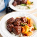 A white plate filled with slow cooker garlic honey meatballs in sauce, steamed rice, and mixed vegetables including carrots, green beans, and squash. Another similar plate is blurred in the background.