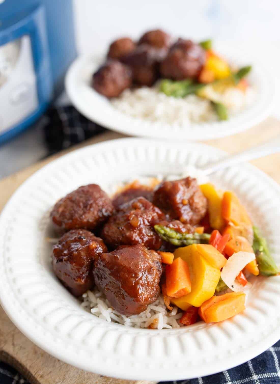A white plate filled with slow cooker garlic honey meatballs in sauce, steamed rice, and mixed vegetables including carrots, green beans, and squash. Another similar plate is blurred in the background.
