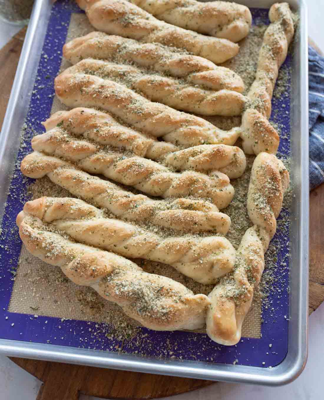 A baking sheet with several twisted breadsticks sprinkled with herbs and seasonings, resting on a silicone baking mat on a wooden surface.
