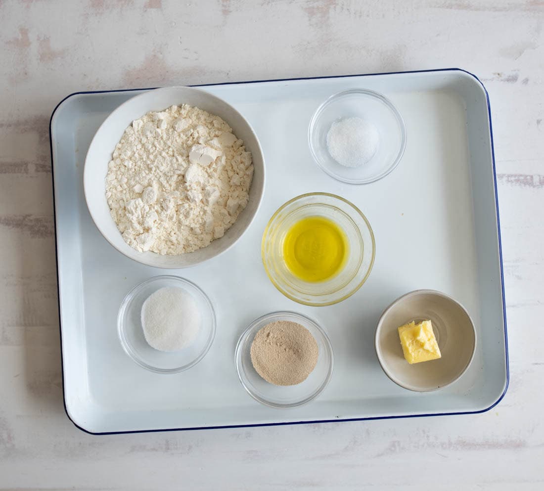 A white tray holds bowls containing flour, sugar, dry yeast, salt, olive oil, a pat of butter, and another white powder, all arranged neatly on a light surface.