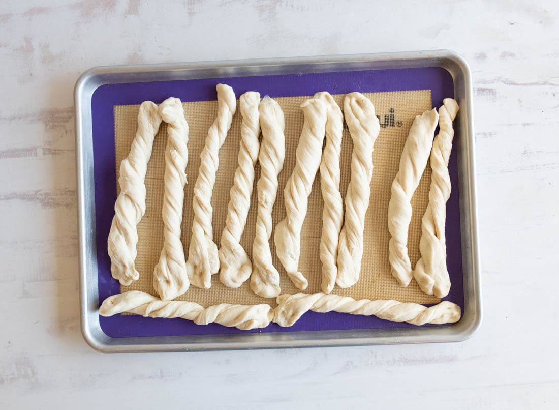 A baking sheet lined with a silicone mat holds several twisted strips of raw dough, evenly spaced and ready for baking.