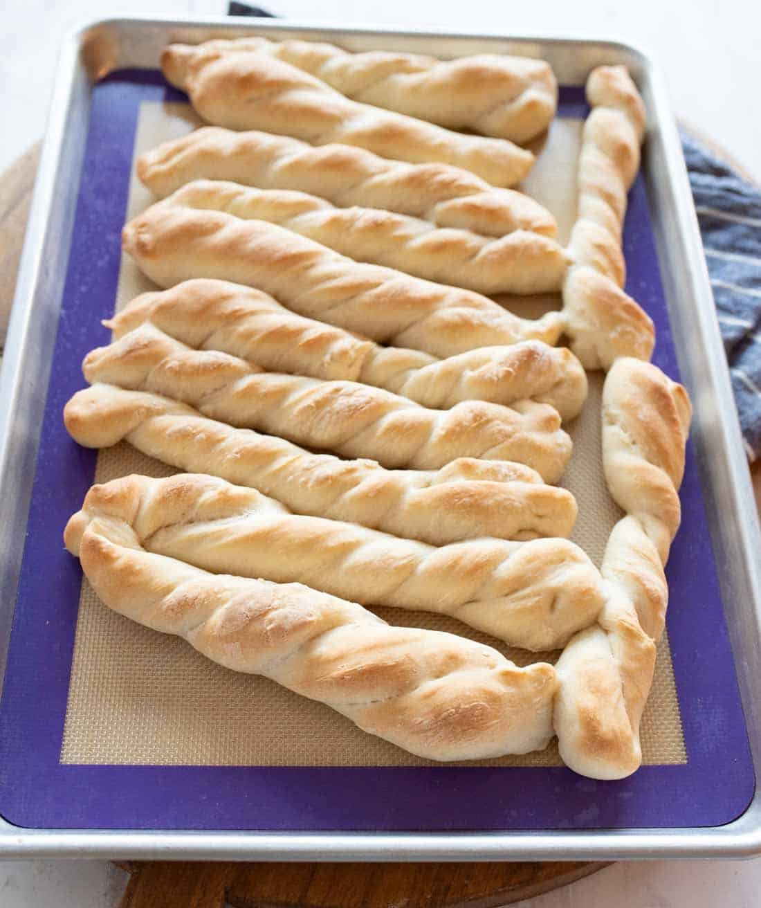 A baking tray lined with a silicone mat holds several golden-brown, twisted breadsticks arranged in rows, fresh out of the oven.