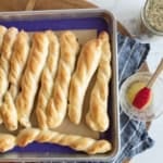 A baking tray with several golden, twisted breadsticks on a blue and tan cloth, next to a jar of seasoning and a bowl of melted butter with a red pastry brush.