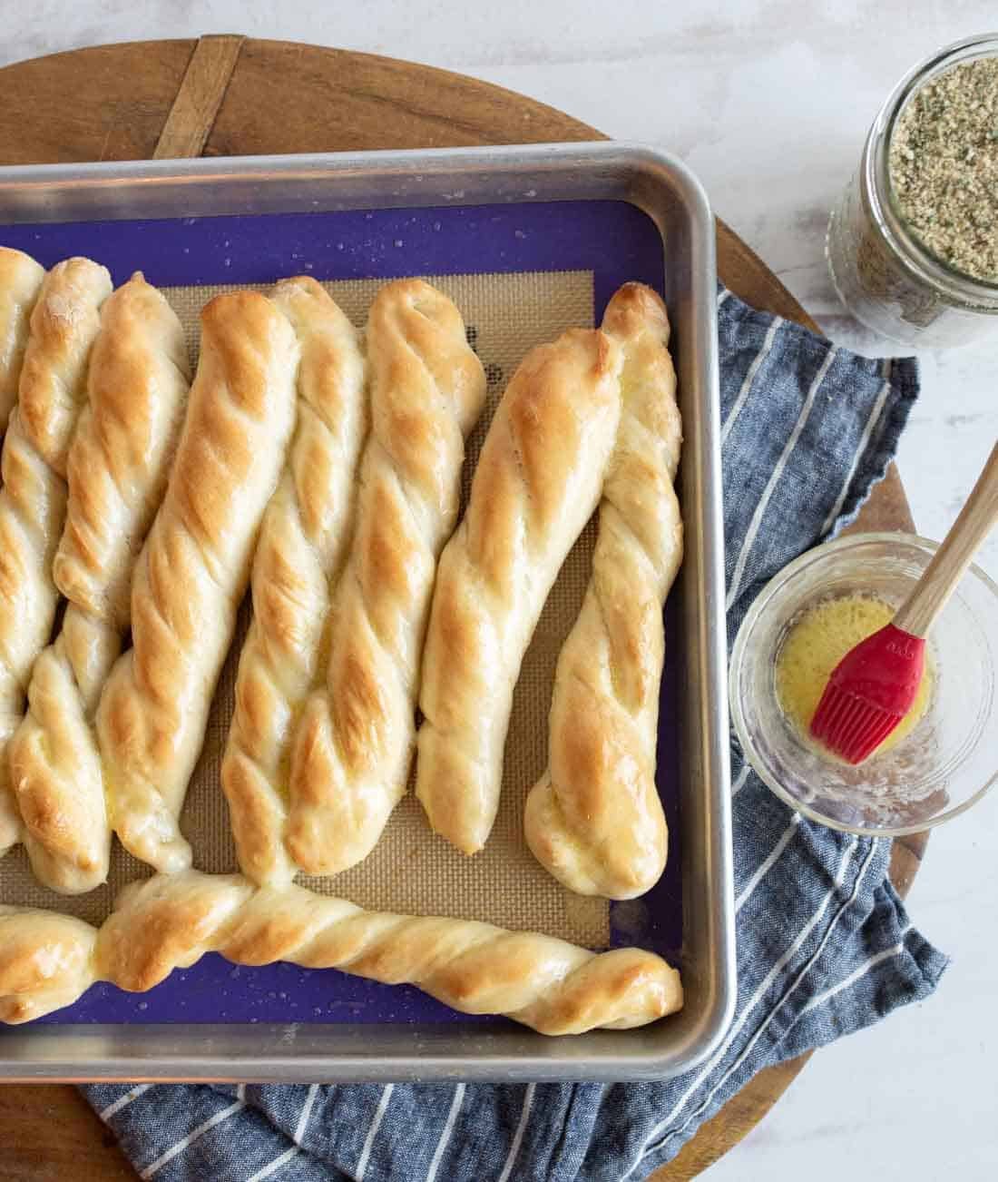 A baking tray with several golden, twisted breadsticks on a blue and tan cloth, next to a jar of seasoning and a bowl of melted butter with a red pastry brush.