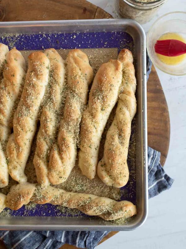 A baking tray with several twisted Italian breadsticks sprinkled with herbs, sitting on a blue cloth, and a jar with a red basting brush and oil nearby.