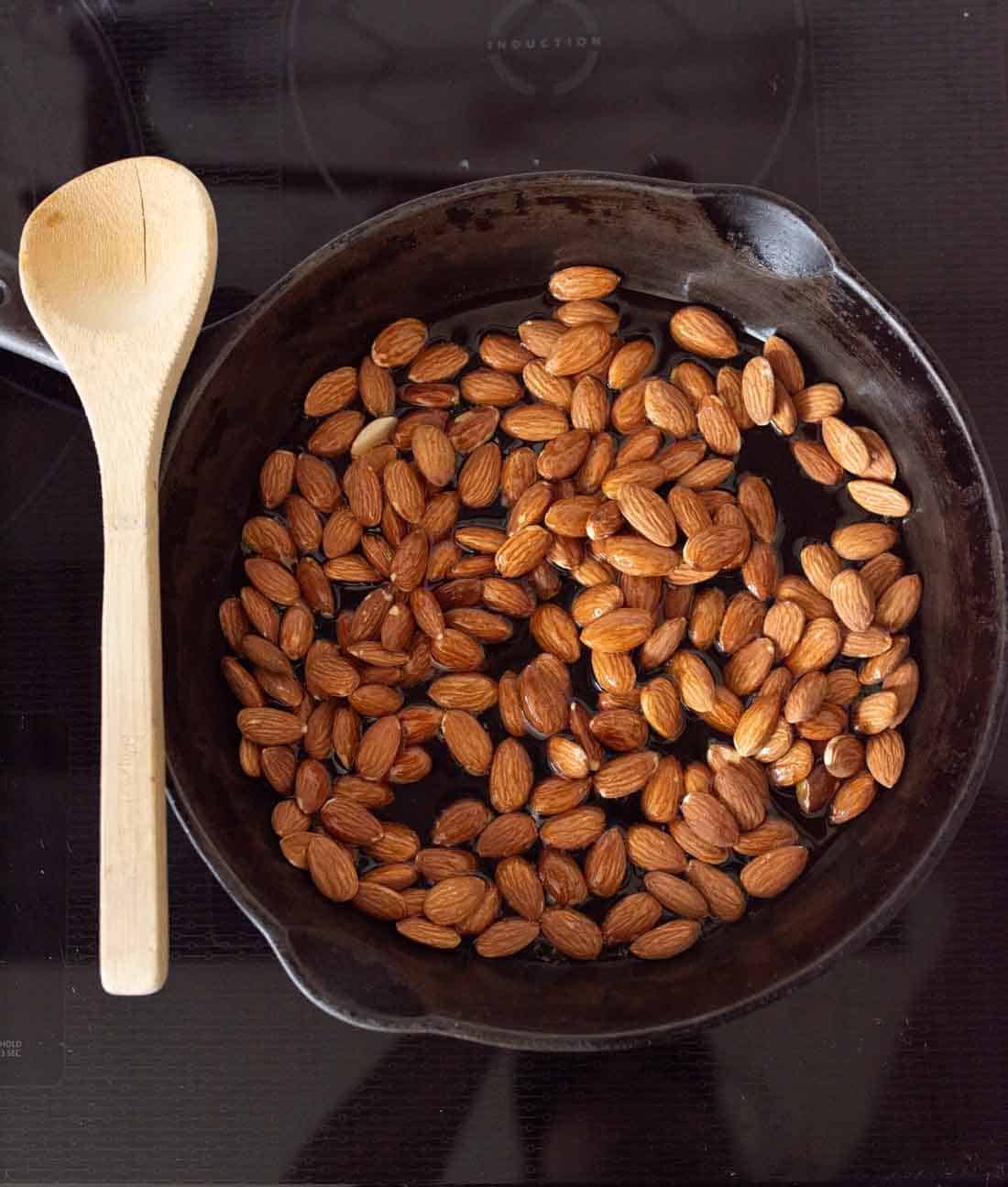 A cast iron skillet filled with raw almonds sits on a stovetop, with a wooden spoon placed next to the skillet.
