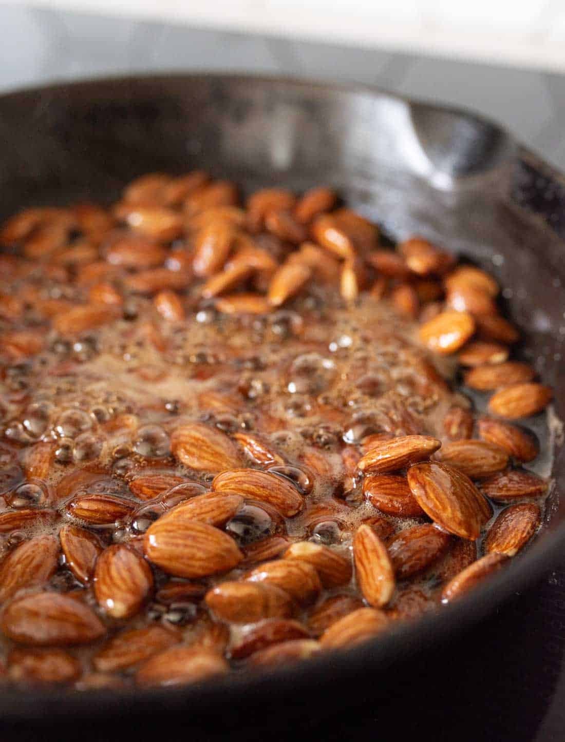 Whole almonds simmer in bubbly, caramelized liquid in a black cast iron skillet on a stovetop.
