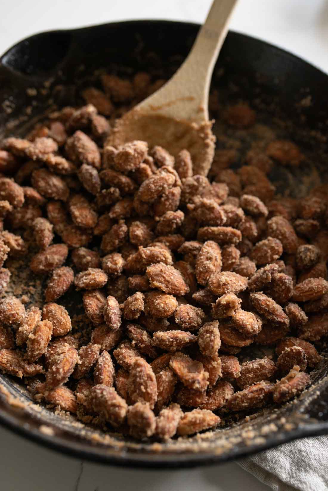 A wooden spoon stirs a pan filled with roasted almonds coated in a crunchy, sugary cinnamon mixture.