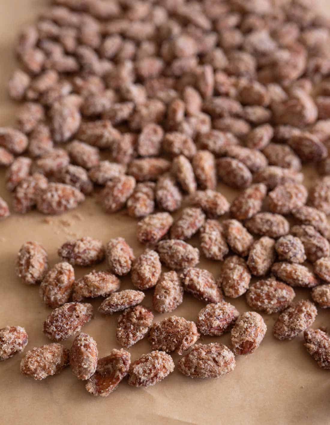 A close-up view of many sugar-coated roasted almonds, crafted using a classic candied almonds recipe, spread out on a sheet of brown parchment paper.