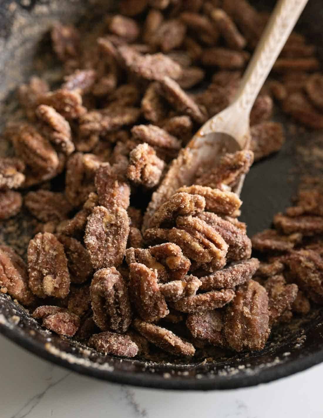 A close-up of candied pecans coated in cinnamon sugar, being stirred with a wooden spoon in a black bowl.