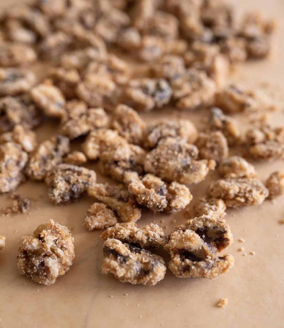 Close-up of clusters of walnuts, coated in a layer of sugar and spices, scattered on a sheet of brown parchment paper from a candied walnuts recipe.