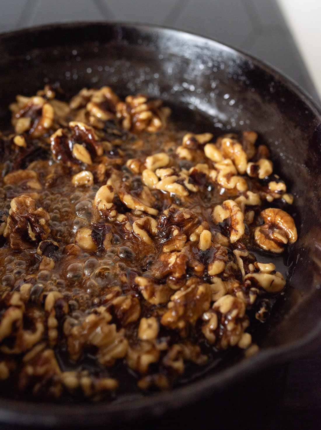 Walnut halves sizzling in hot oil in a dark skillet, with small bubbles forming around the nuts as they cook. The background is blurred and dark, highlighting the cooking process.