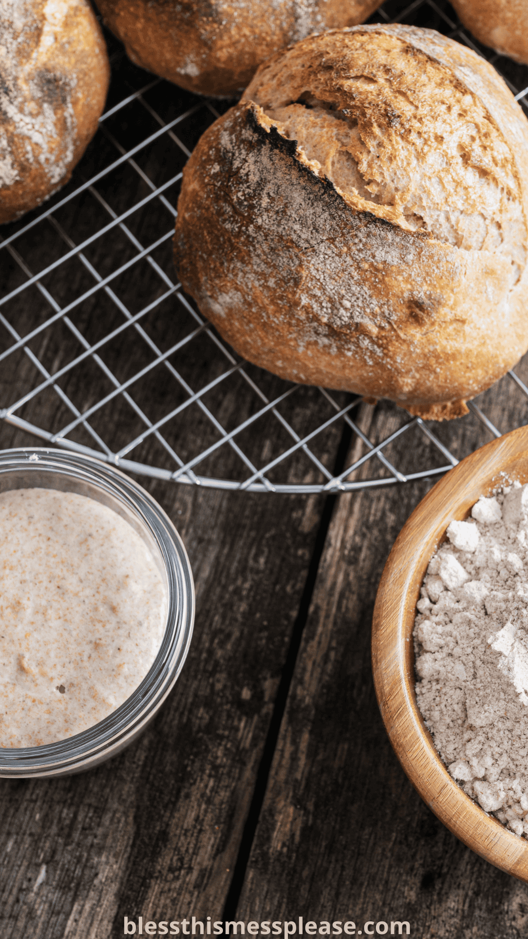 Crusty round loaves of bread on a cooling rack, a jar of sourdough starter, and a wooden bowl of flour rest on a rustic wooden surface—perfect for exploring the differences between instant yeast vs active dry yeast in baking.
