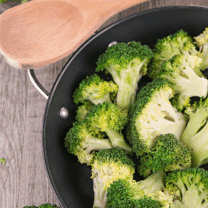 A frying pan filled with fresh broccoli florets sits on a wooden surface next to a wooden spoon, with more broccoli for how to preserve broccoli and a website label blessthismessplease.com visible.