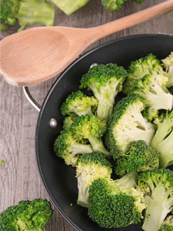 A frying pan filled with fresh broccoli florets sits on a wooden surface next to a wooden spoon, with more broccoli for how to preserve broccoli and a website label blessthismessplease.com visible.