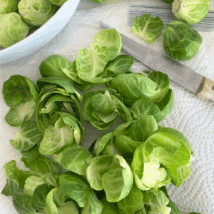 Brussels sprouts being prepped on a paper towel showing how to preserve brussels sprouts, with some whole and some trimmed or halved, a knife and a white bowl nearby. A green label at the bottom reads blessthismessplease.com.