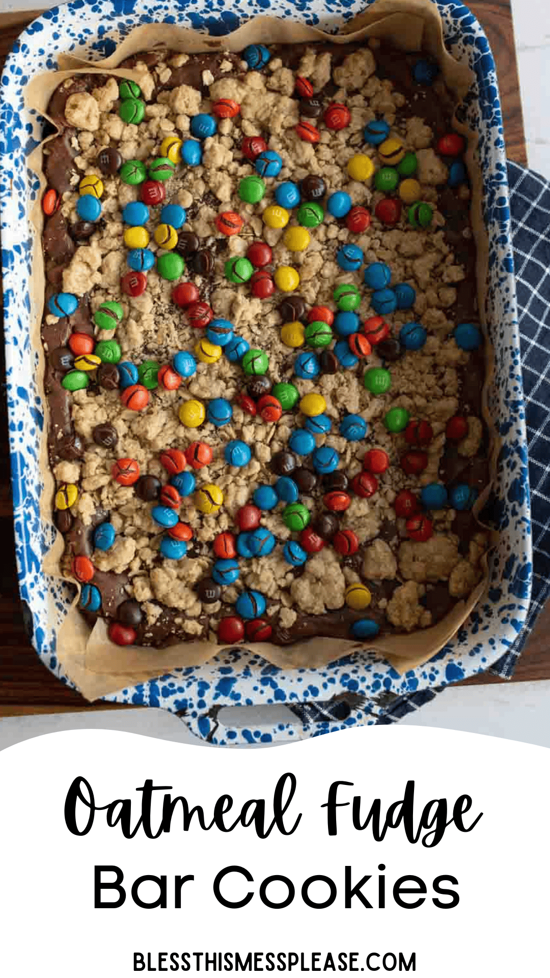 A rectangular baking dish lined with parchment paper holds oatmeal fudge bars topped with colorful M&M candies and oatmeal crumbs. The dish sits on a blue-patterned cloth.