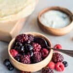 A wooden bowl filled with mixed berries, including raspberries, blackberries, and blueberries, with a spoon inside. Nearby are another bowl of mixed berry compote and a stack of crepes on a wooden board.