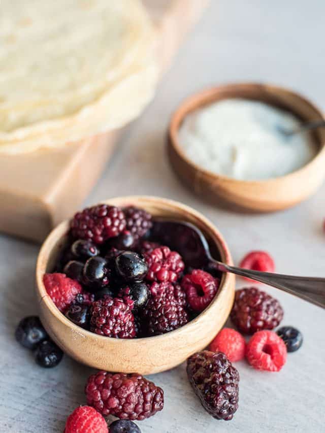 A wooden bowl filled with mixed berries, including raspberries, blackberries, and blueberries, with a spoon inside. Nearby are another bowl of mixed berry compote and a stack of crepes on a wooden board.