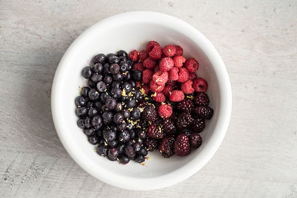 A white bowl containing three groups of fresh berries—perfect for a berry compote—features blueberries, raspberries, and blackberries arranged side by side on a light-colored surface.