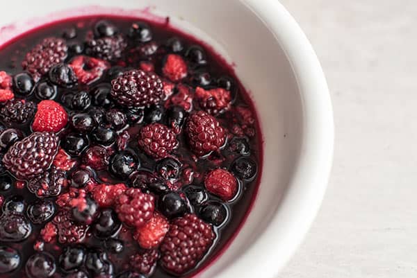 A close-up of a white bowl filled with mixed berries, including blackberries, blueberries, and raspberries, served in a rich berry compote sauce on a light surface.