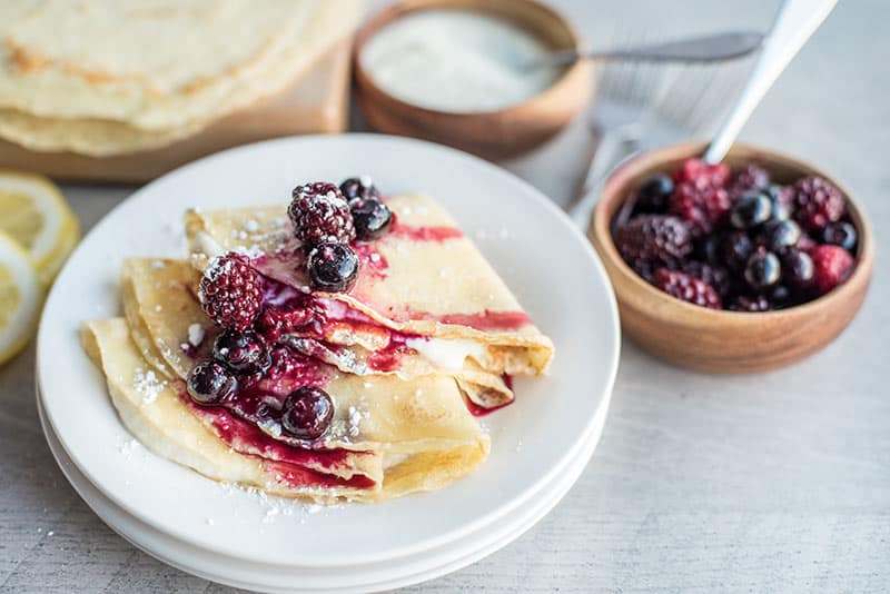 A plate of folded crepes topped with powdered sugar and mixed berries, served alongside a vibrant berry compote, with a bowl of cream and stacked crepes in the background on a light table.