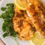 Two seasoned, blackened chicken breasts on a white plate, garnished with fresh parsley and lemon slices.