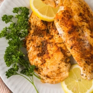 Two seasoned, blackened chicken breasts on a white plate, garnished with fresh parsley and lemon slices.