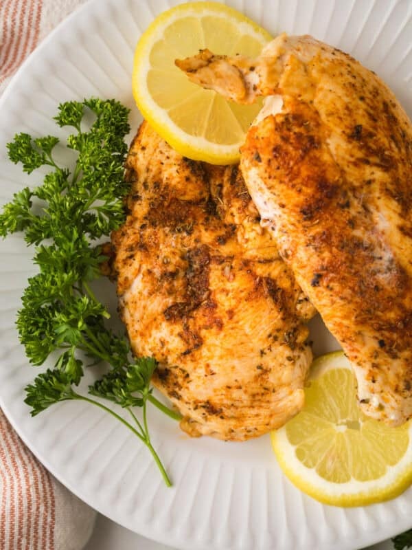 Two seasoned, blackened chicken breasts on a white plate, garnished with fresh parsley and lemon slices.