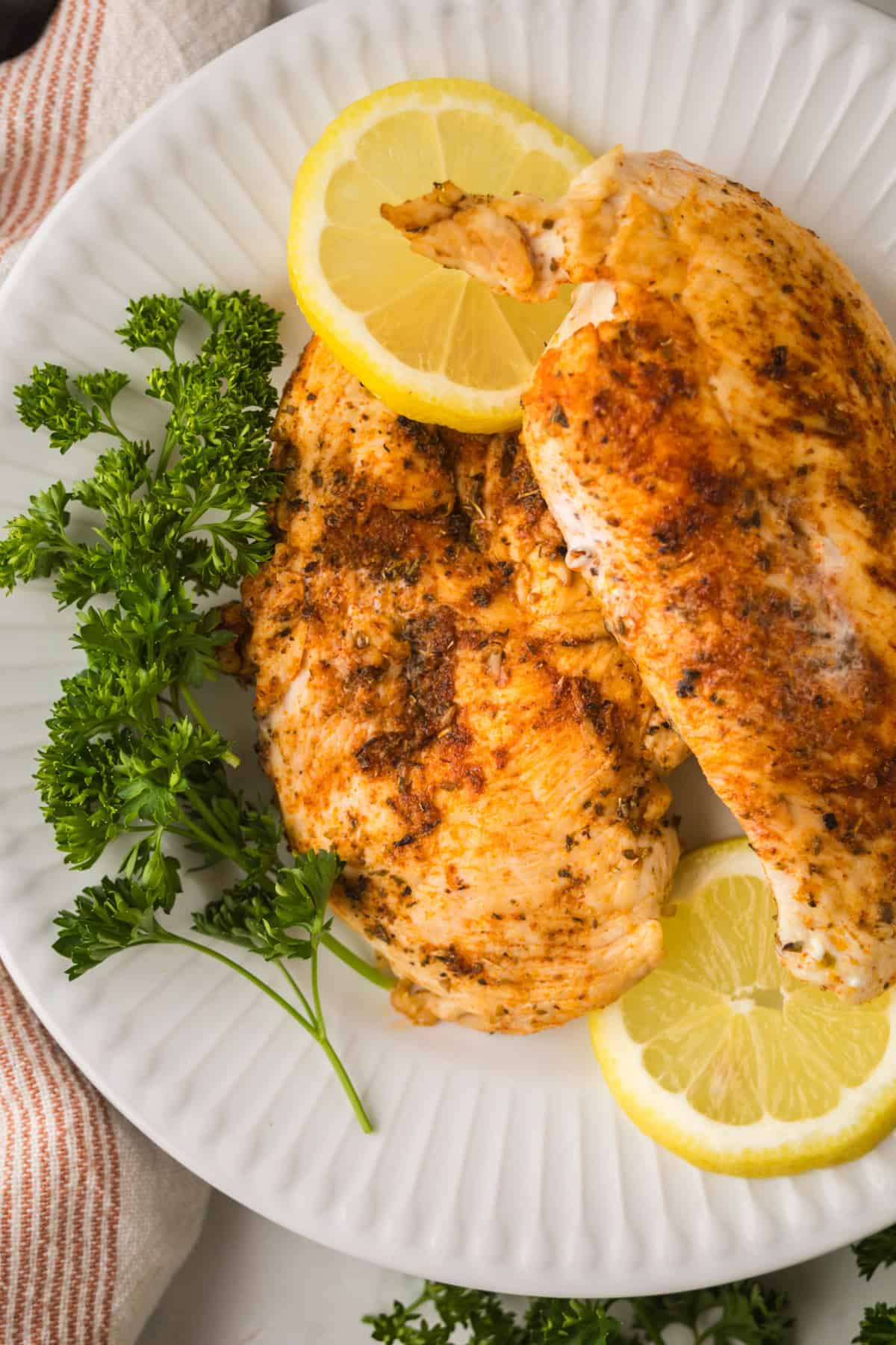 Two seasoned, blackened chicken breasts on a white plate, garnished with fresh parsley and lemon slices.