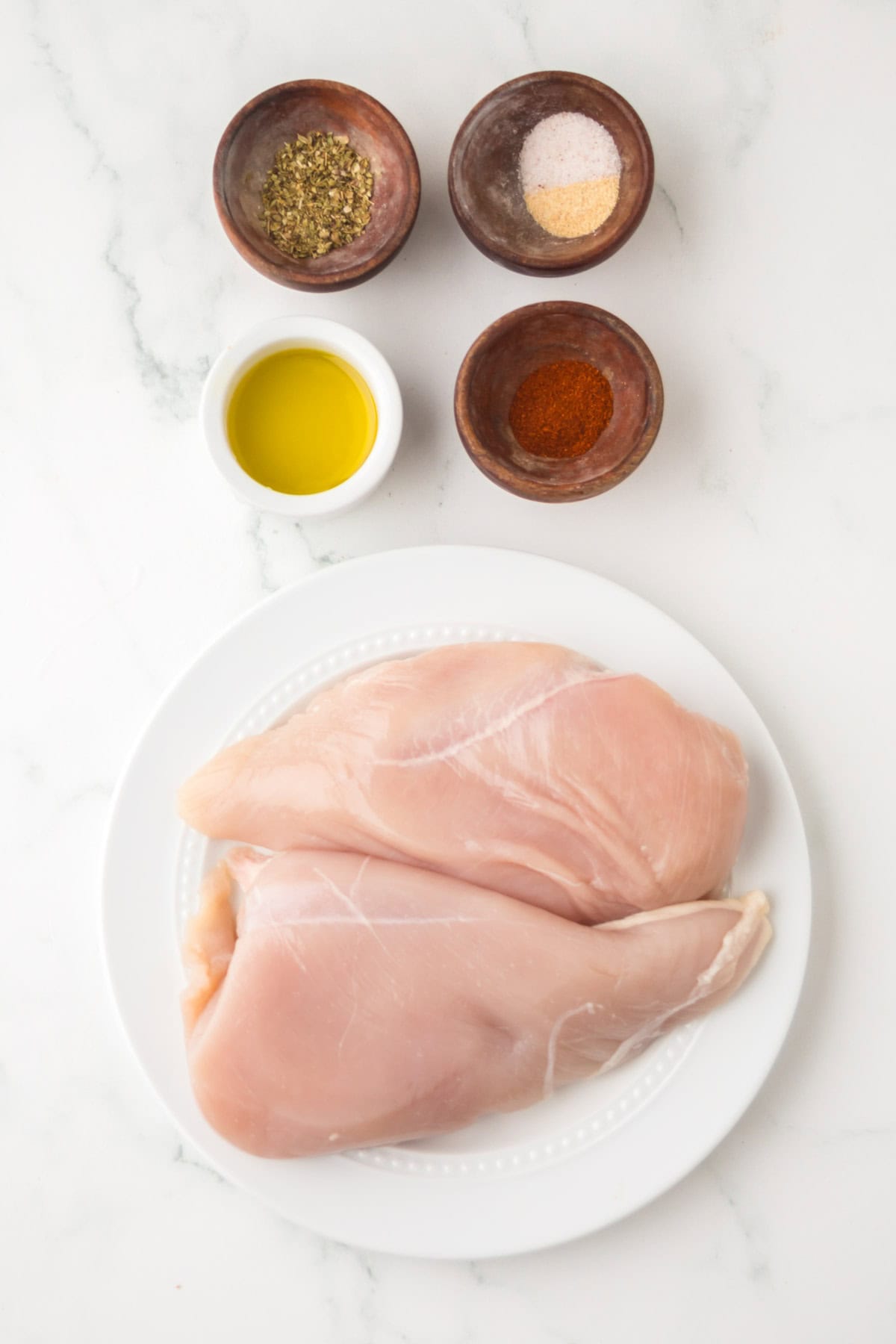 Two raw chicken breasts on a white plate, surrounded by small bowls of olive oil, dried herbs, blackened chicken seasoning, spices, and salt, all arranged on a white marble surface.