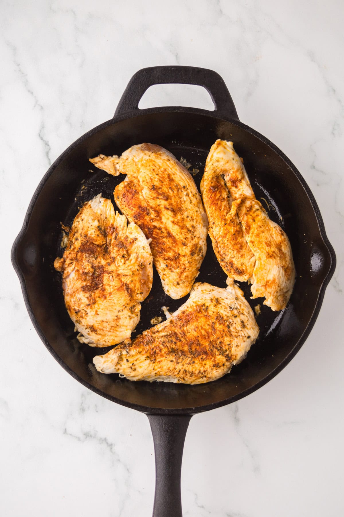 Four seasoned blackened chicken breasts cooking in a black cast iron skillet on a white marble surface.