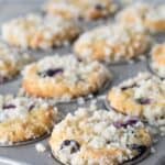 A close-up of freshly baked blueberry streusel muffins in a metal tin, crumbly topping glistening, with a few muffins slightly out of focus in the background.