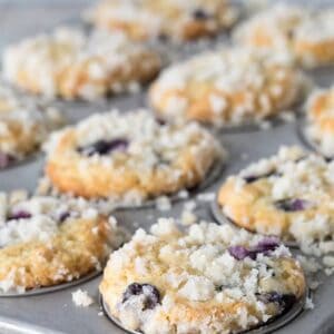 A close-up of freshly baked blueberry streusel muffins in a metal tin, crumbly topping glistening, with a few muffins slightly out of focus in the background.