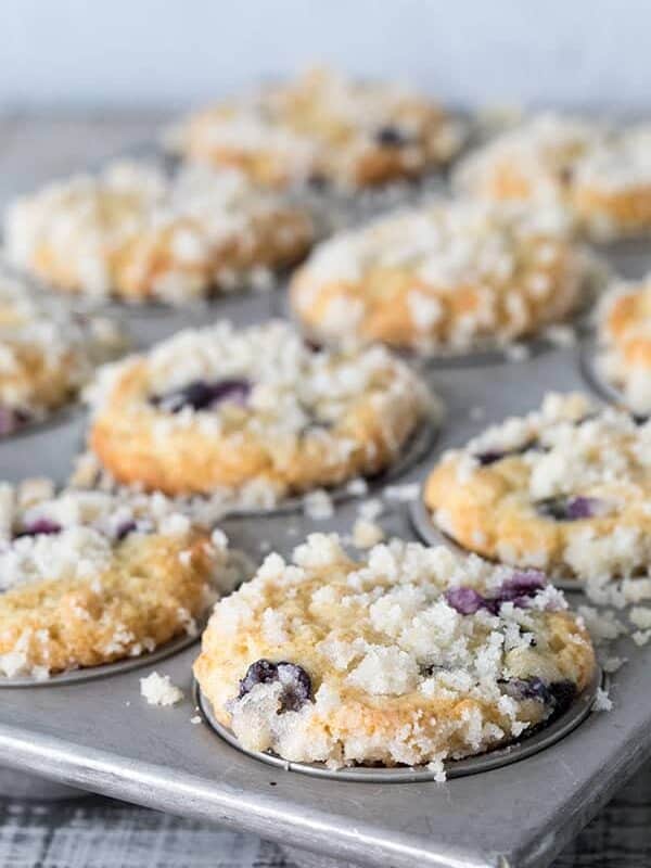 A close-up of freshly baked blueberry streusel muffins in a metal tin, crumbly topping glistening, with a few muffins slightly out of focus in the background.