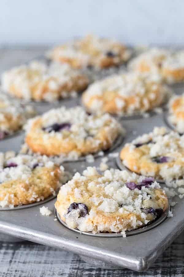 A close-up of freshly baked blueberry streusel muffins in a metal tin, crumbly topping glistening, with a few muffins slightly out of focus in the background.