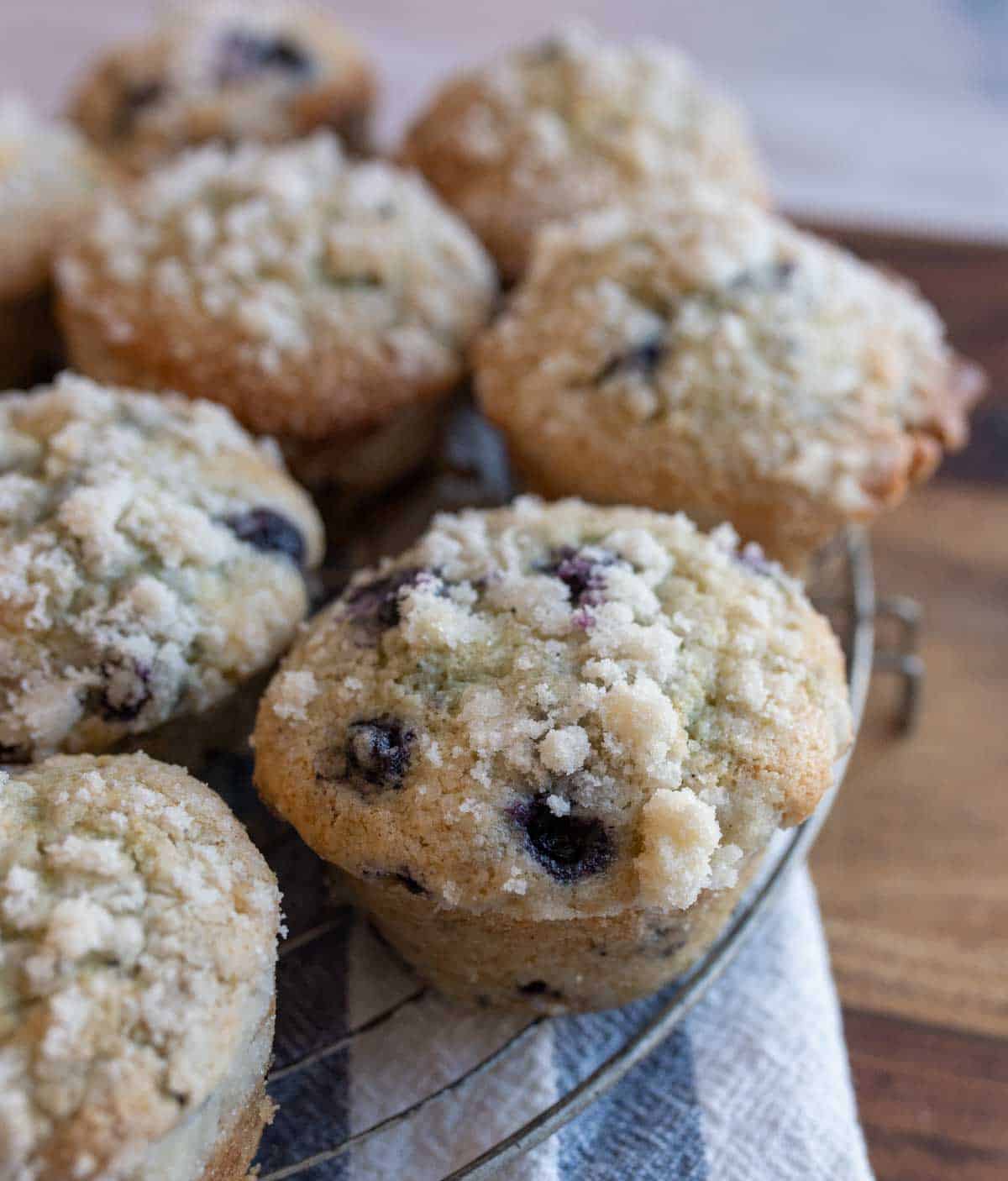 A close-up of blueberry streusel muffins with crumbly topping, arranged on a wire rack over a striped cloth. The muffins are golden brown with visible blueberries and glistening sugar on top.