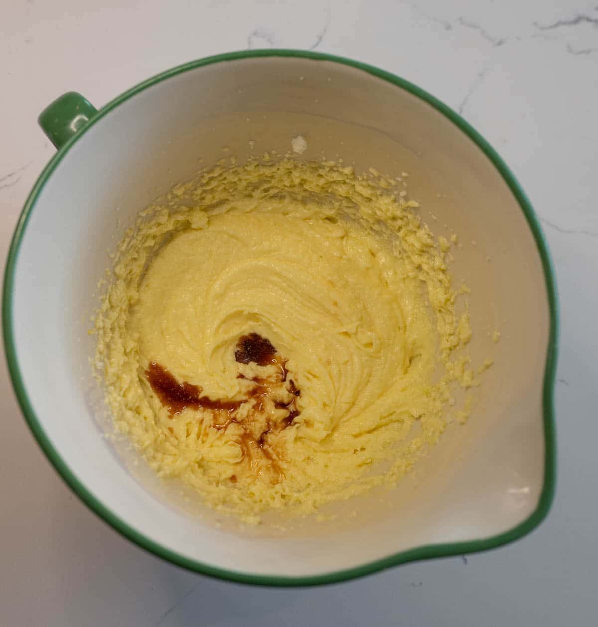 A green mixing bowl containing creamed butter and sugar, with a small amount of vanilla extract added on top, sits on a white countertop—perfect for starting your blueberry streusel muffins.